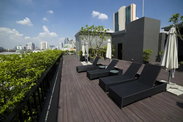 A rooftop terrace with black wicker sun loungers, closed white umbrellas and potted plants, with high-rise buildings visible under a blue sky.