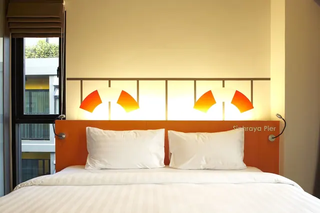 A double bed with white pillows and linen sits against an orange headboard with wall lamps, and a window on the left shows a neighbouring building and trees.