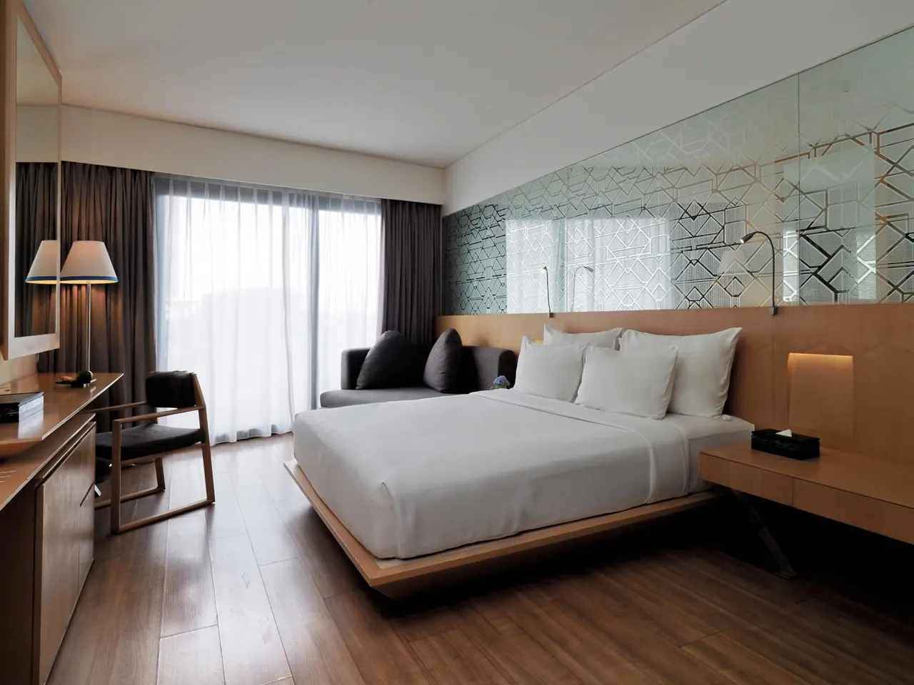 A bedroom with a king bed, a grey sofa, a wooden desk and chair, and sheer-curtained floor-to-ceiling windows.