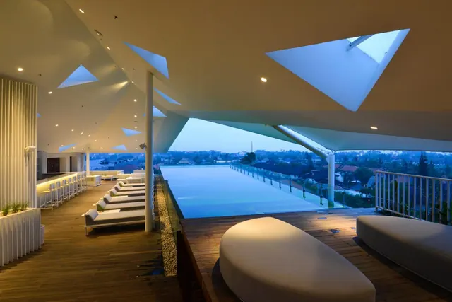 A rooftop infinity pool with white sun loungers and a sculptural ceiling of triangular skylights, photographed at dusk.