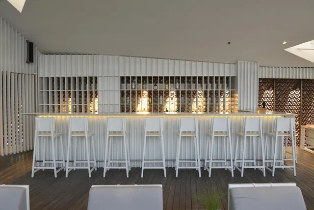 A white slatted bar with backlit shelves of bottles, white bar stools along the counter, and white lounge chairs in the foreground on a timber deck.