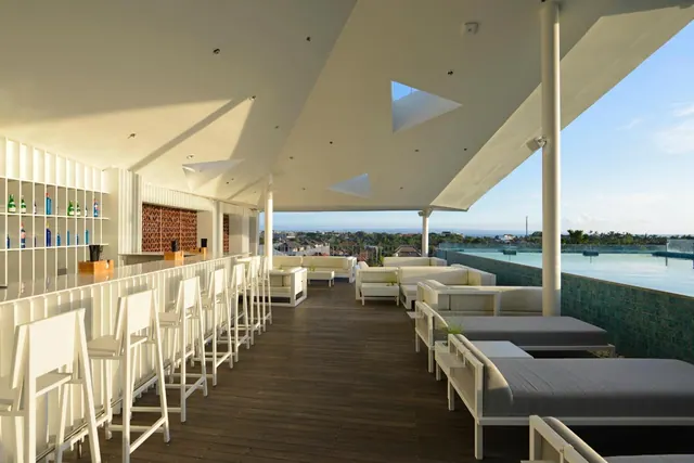 A rooftop bar with a white counter, high stools and white sofas and daybeds on a timber deck, with an infinity pool and distant sea to the right.