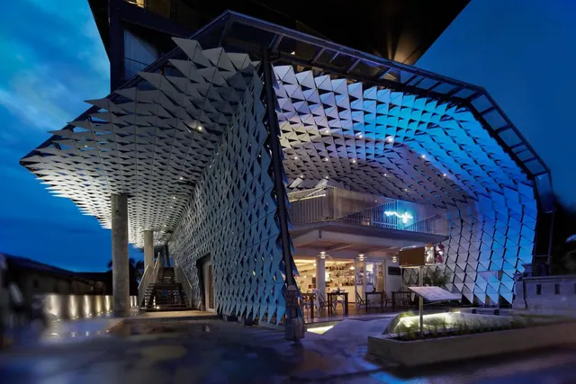 A building exterior at dusk with a large folded geometric latticed facade lit in blue, with a staircase and an illuminated ground-floor interior visible beneath.