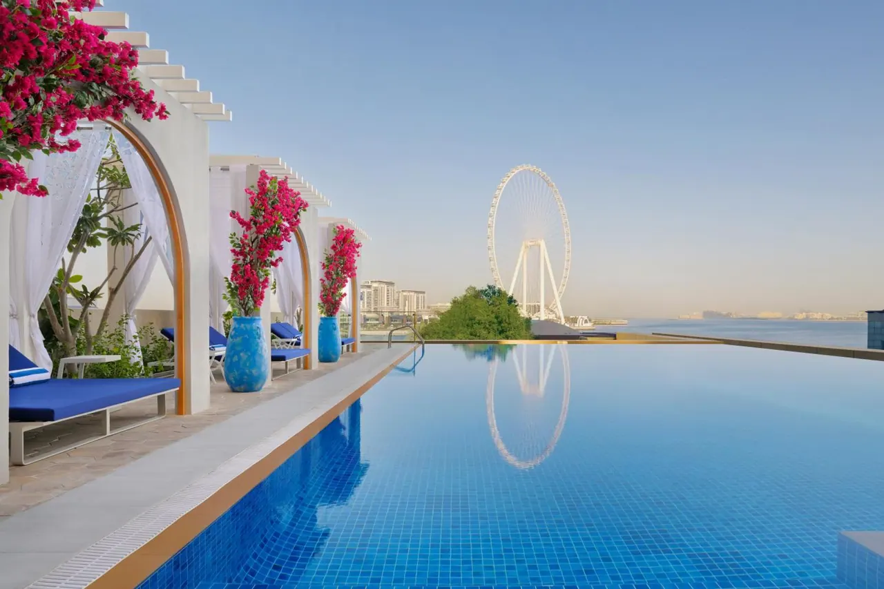 An infinity-edge pool runs beside white cabanas with blue loungers, with the Ain Dubai ferris wheel and the sea visible in the distance.