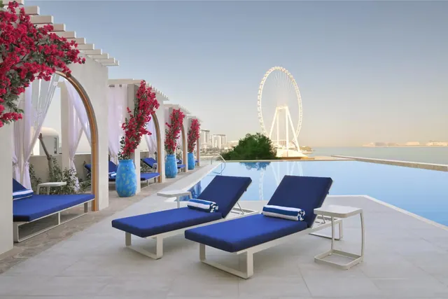 Two blue sun loungers with white towels sit beside an infinity pool, with the Ain Dubai ferris wheel and the sea visible beyond.