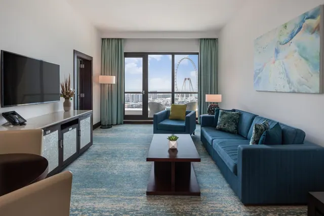 A hotel living room with a blue sofa, a wall-mounted television, and glass doors to a balcony with chairs, with the Ain Dubai ferris wheel visible outside.