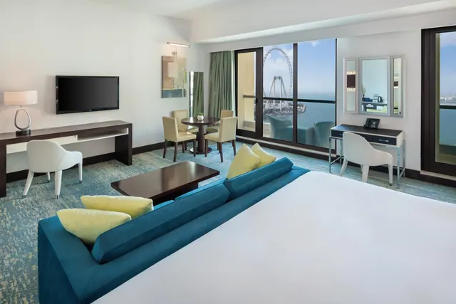 A hotel bedroom with a large white bed, a blue sofa, a dining table, a desk and a TV, with floor-to-ceiling windows and a balcony view of the Ain Dubai observation wheel and the sea.