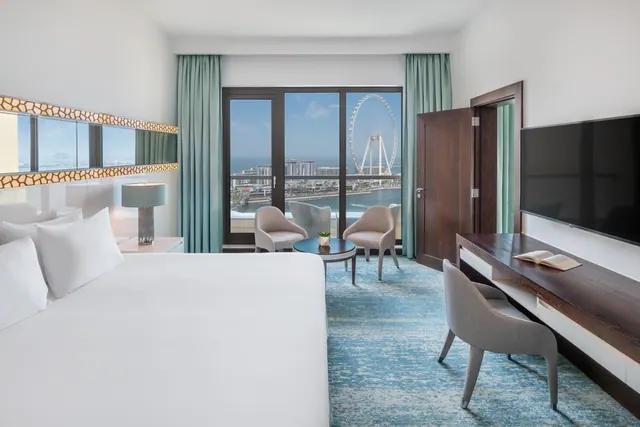 A bedroom with a large white bed, armchairs by a glass door, and a view of the Ain Dubai ferris wheel and the sea.