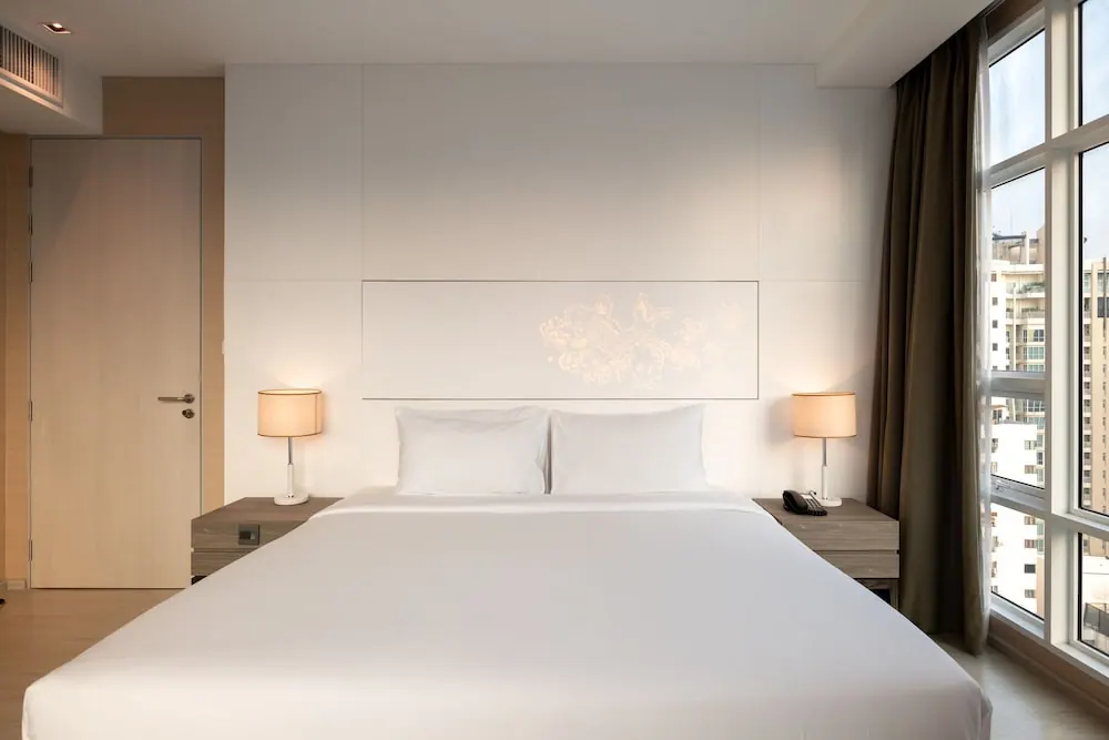 A bedroom with a made king bed, two bedside lamps, and floor-to-ceiling windows looking out onto surrounding buildings.
