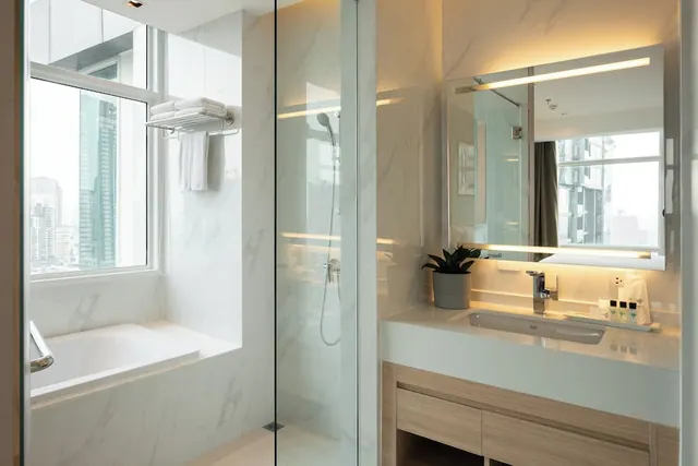 A bathroom with a bathtub beside a window overlooking high-rise buildings, a glass-walled walk-in shower, and a single basin vanity beneath a backlit mirror.