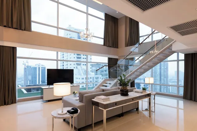 A double-height living room with a grey sofa, a television on a white media unit, and floor-to-ceiling windows looking out over high-rise buildings, with a glass-railed staircase on the right.