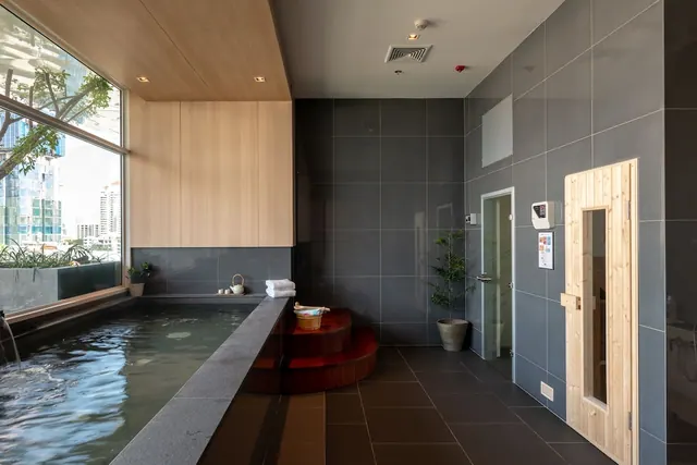 A spa room with a sunken hot tub with wooden steps, a wooden sauna door, and a large window showing trees and city buildings outside.