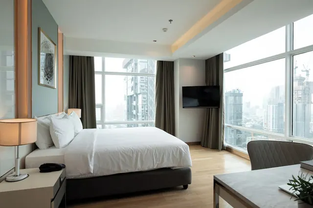 A bedroom with a made bed, a wall-mounted television, a desk and floor-to-ceiling windows overlooking high-rise buildings.