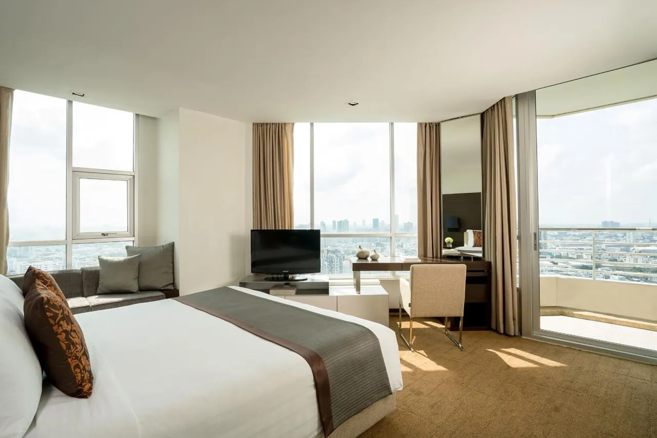 A bedroom with a large white bed, a seating area by the windows, a desk and television, and sliding doors to a balcony overlooking the city.