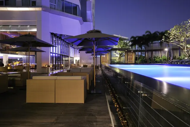 An outdoor pool with a glass edge at night, beside wicker sofas under umbrellas on a timber deck.