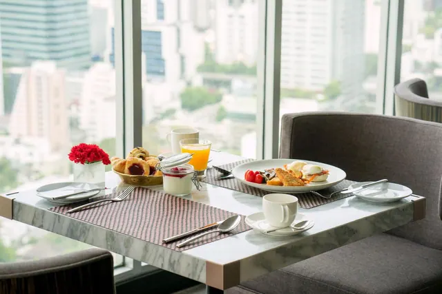 A breakfast table with plated food, orange juice, pastries and a red flower arrangement sits beside floor-to-ceiling windows overlooking city buildings.