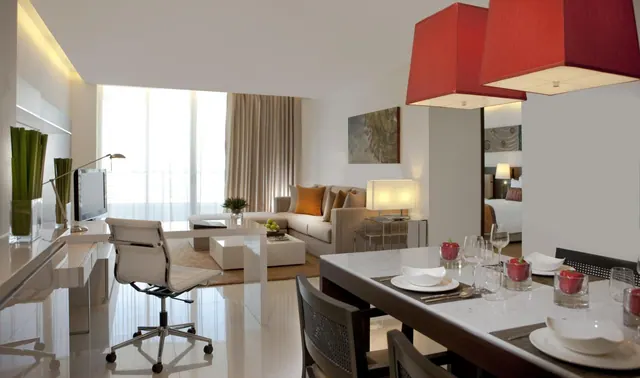 A staged suite living area with a white desk and chair, a sofa with orange cushions, a dining table under red pendant lamps, and a bed visible through a doorway.