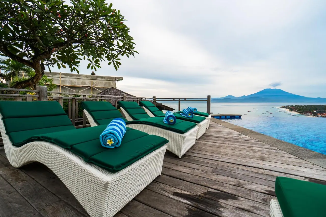 Green-cushioned white wicker sun loungers with rolled blue towels sit on a wooden deck beside a pool that overlooks the sea and a distant volcanic mountain.