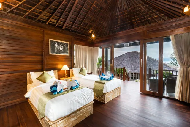 A bedroom with two beds dressed with towel animals, wooden walls and ceiling, and glass doors opening onto a balcony with a view of rooftops.