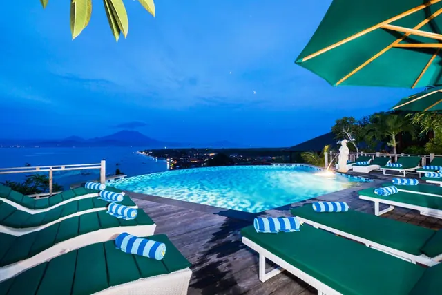 An infinity pool at dusk with green-cushioned sun loungers and a green umbrella, overlooking the sea and a distant mountain.