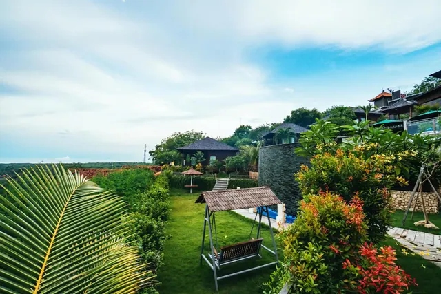 A grassy garden with a covered swing seat, tropical shrubs and a palm frond in the foreground, with villa roofs on the hillside behind.