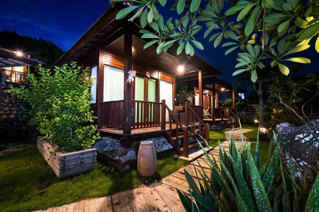 A wooden bungalow with a raised veranda, railings and steps is lit at night, with a wooden walkway and planted garden in front.