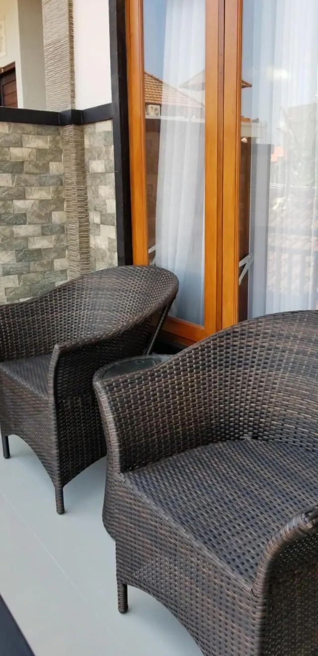 Two woven rattan armchairs and a small side table sit on a tiled balcony beside a wooden-framed glass door with sheer curtains.