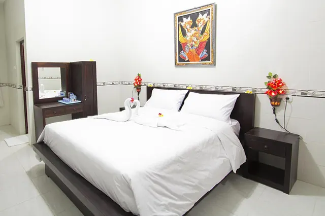 A king bed with white linen and a dark wooden headboard sits beside a dressing table with a mirror, under a framed painting of Balinese dancers.