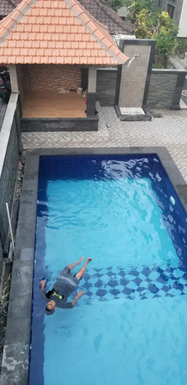 A person floats in a rectangular blue-tiled pool beside a tiled gazebo with a terracotta roof and a stone outdoor shower.