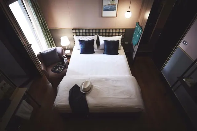 A double bed with white linen and navy cushions sits beside an armchair and a window in a dim bedroom, with a hat and jacket laid on the covers.