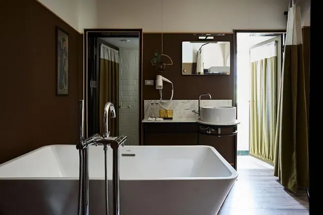 A freestanding bathtub with a floor-mounted tap sits in a bathroom beside a curtained shower area, a vessel sink on a marble counter, and a wall-mounted hair dryer.