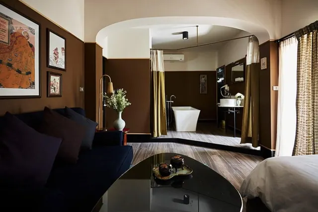 A room with a navy sofa, framed artwork and a black glass coffee table, with an arched opening onto a bathroom containing a freestanding bathtub.