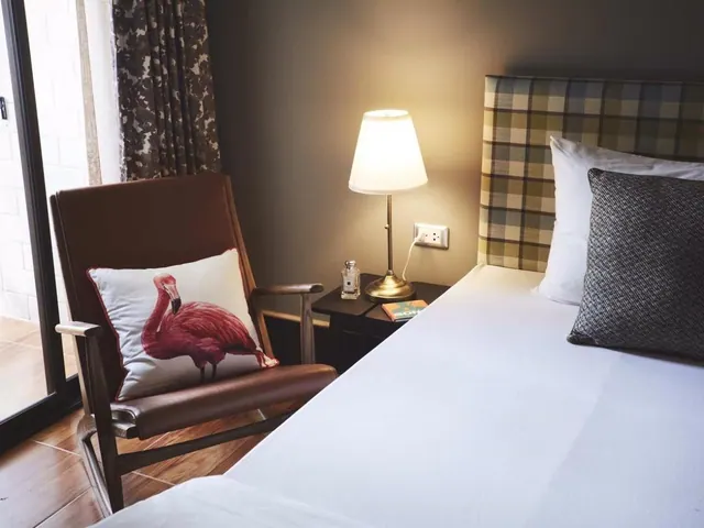 A bedroom with a made white bed, plaid headboard, a leather armchair with a flamingo cushion, and a lit bedside lamp beside a glass sliding door.