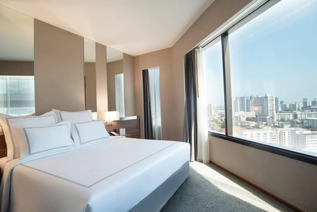 A bedroom with a made-up king bed with white linens beside a floor-to-ceiling window overlooking high-rise buildings.