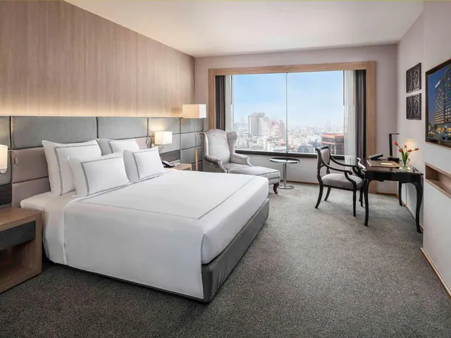 A king bed with white linen faces a large window onto city buildings, with an armchair, a desk with chair and a wall-mounted television in the room.
