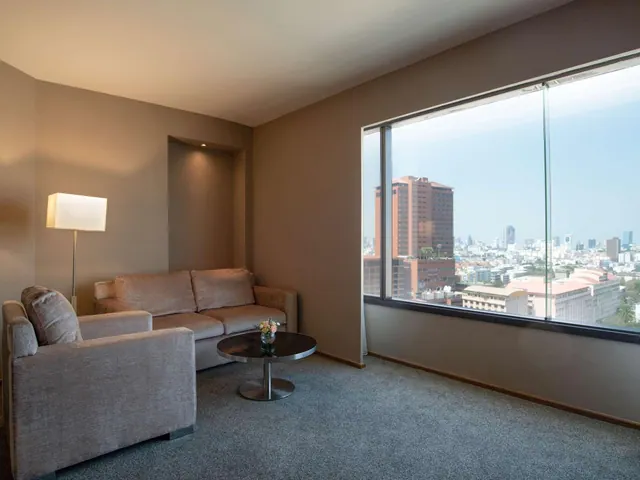 A living area with a beige sofa, armchair, floor lamp and round coffee table, beside a large window overlooking a city skyline.