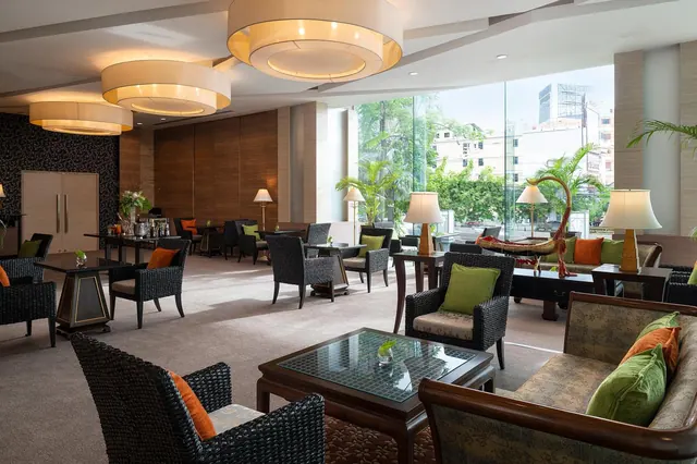 A lounge seating area with dark wicker armchairs, green and orange cushions, drum pendant lights and floor-to-ceiling windows looking onto palm trees and low buildings.