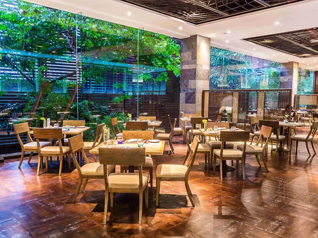 A restaurant dining room with wooden tables and cushioned chairs, with floor-to-ceiling glass windows looking onto leafy trees outside.