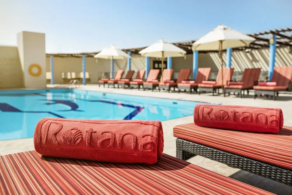 Two rolled red towels embossed with the Rotana logo lie on striped loungers beside a rectangular swimming pool, with white umbrellas and sun loungers under a clear sky.