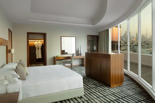 A bedroom with a made king bed, a desk and chair, a wardrobe with robes, and floor-to-ceiling windows overlooking a city skyline at dusk.