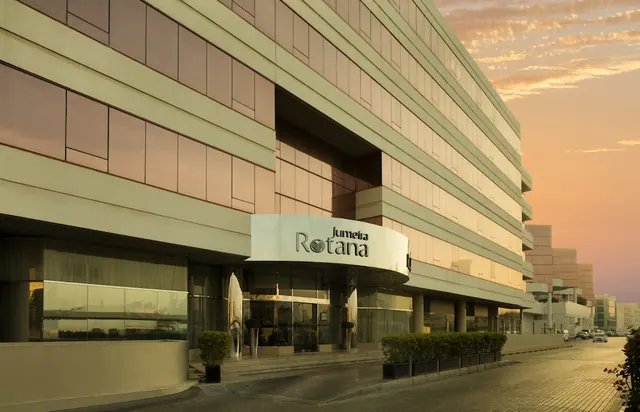 Exterior of the Jumeira Rotana hotel at sunset, showing a curved entrance canopy with the hotel sign, planters at the entrance, and cars on the road beside the building.