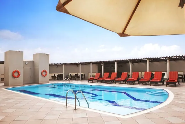An outdoor swimming pool with red sun loungers under a pergola and a beige parasol, with life rings on a wall beside the pool.