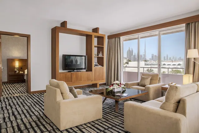 A living area with beige armchairs and a sofa around a glass coffee table, a wall-mounted TV in a wooden unit, and a view through the windows of the Dubai skyline with Burj Khalifa.