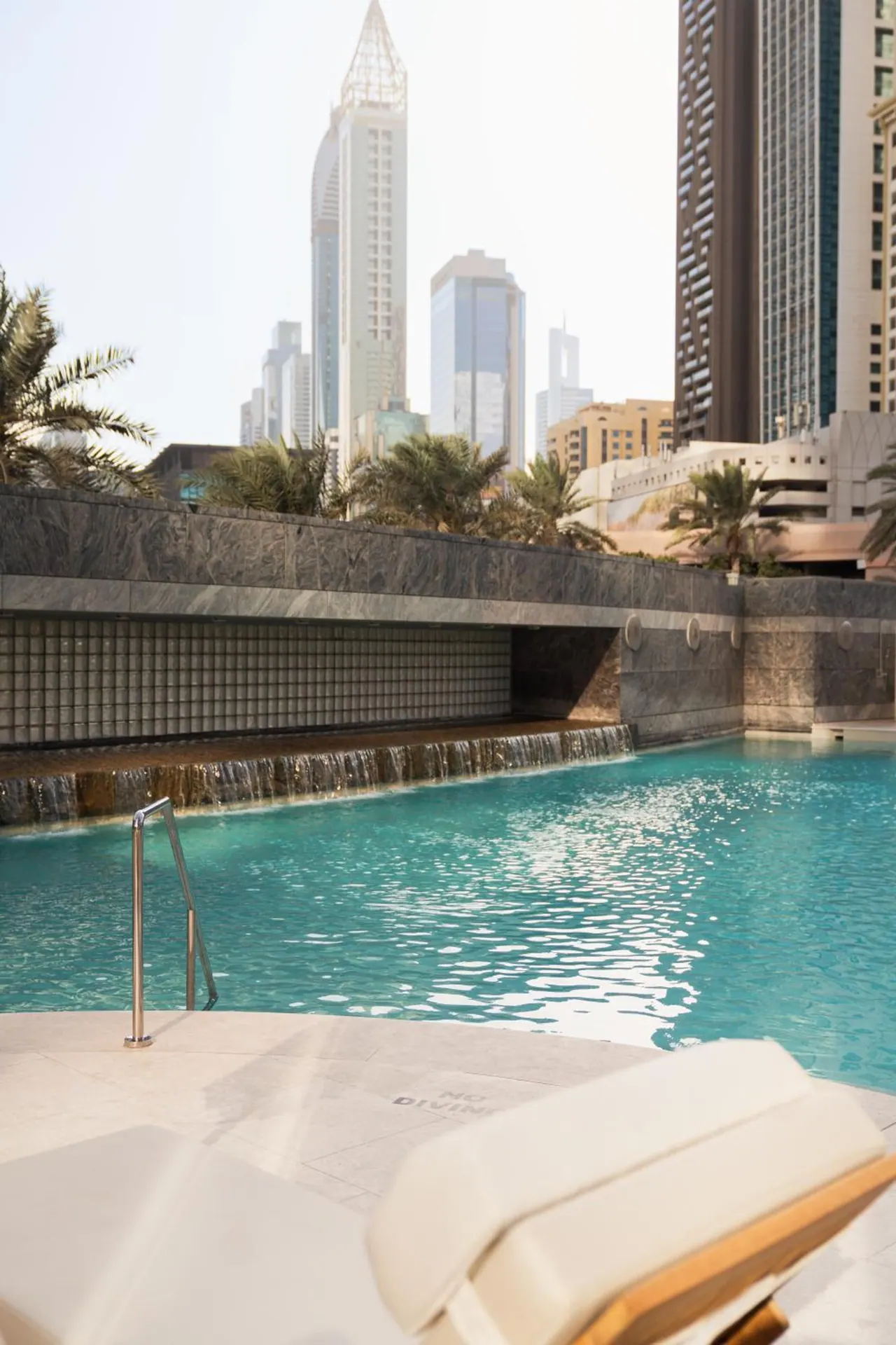 A turquoise pool with a waterfall spilling from a stone wall, palm trees and high-rise towers behind, and a cream sun lounger in the foreground.