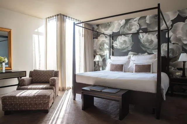 A bedroom with a four-poster king bed, a patterned armchair and ottoman, and floral wallpaper beside a window with sheer curtains.