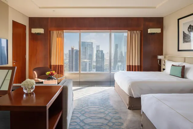 A hotel bedroom with a large bed, a seating area and floor-to-ceiling windows looking out over a city skyline of tall towers.
