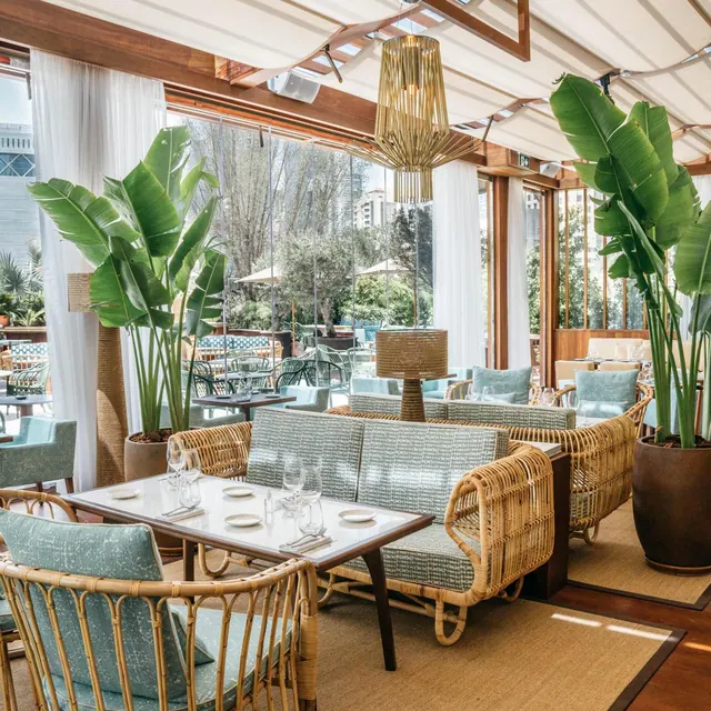 A covered restaurant dining area with rattan sofas and armchairs, set tables, large potted banana plants, and glass walls looking out onto trees and an outdoor terrace.