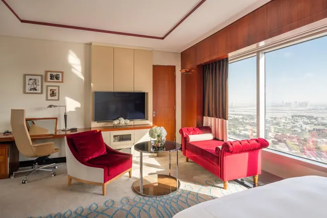 A hotel room seating area with a red armchair, a red chaise, a wall-mounted television, a desk, and large windows overlooking distant city buildings.