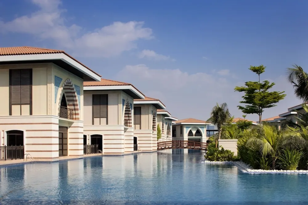 A large pool runs alongside a row of cream-coloured villas with terracotta roofs, with palm trees and a wooden footbridge on the right.