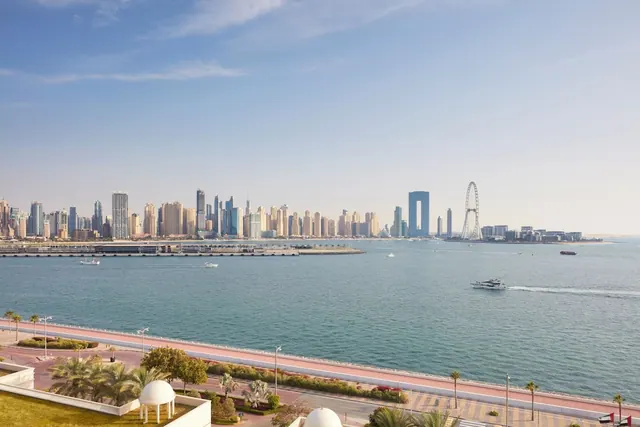 Elevated view across the water toward a high-rise city skyline and the Ain Dubai observation wheel, with a coastal road, palm trees and white domed structures in the foreground.